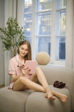 Beauty blogger sits on a couch with her legs crossed while using a tablet. She talks about the