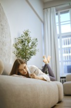 A young woman is lying on a sofa, writing in a notebook with a smile. The room has large windows