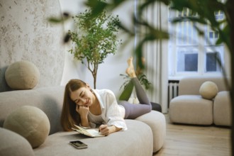 A young woman lies on her stomach on a couch in a modern living room. She is writing in a notebook