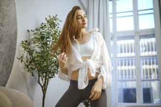 A woman stands indoors by a window with natural light streaming in. She has long hair and wears a