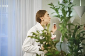 Cute woman in white bathrobe stands in a bathroom with cotton sponge in hand surrounded by green