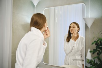 A woman stands in front of a wall mirror in a bathroom. She is washing off makeup from her face