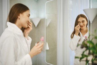 A young woman stands in front of a mirror in the bathroom. She is wearing a bathrobe and removes