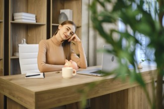 Dreamy woman sitting at a wooden desk in an office. She rests her head on her hand while looking at