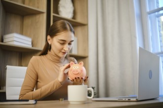 A woman sits at a desk in a home office and smiles while looking at a piggy bank. She is engaging
