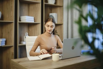 A woman is seated at a wooden desk with a laptop open in front of her. She is writing notes in a
