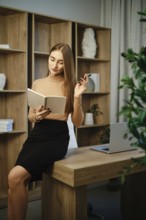 A woman sits on a wooden desk in an office. She is writing in a notebook while holding a pen. A