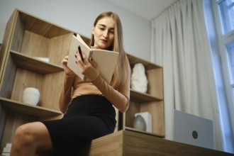 A woman sits on a wooden desk with a notebook in hand. She is focused on reading while surrounded