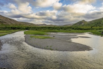 A calm river snakes through a green landscape under a cloudy sky with mountains in the background