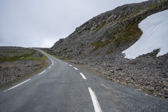 Lonely road snakes through rocky mountain landscape with snow under cloudy sky, Båtsfjord,