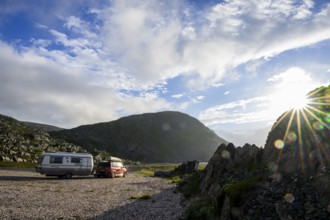 A car with roof box with old Eriba Touring Troll caravan at sunset next to rocks and a mountain in