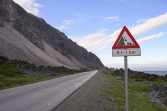 An empty coastal road with a danger sign in front of a mountain range under a blue sky in the north