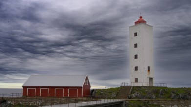 White lighthouse next to a red wooden building against a cloudy sky at the sea Kjølnes Fyr, small