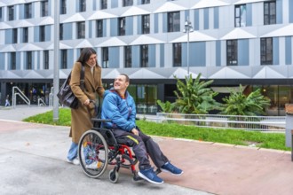 Happy woman caregiver pushing smiling adult man in a wheelchair on an accessible ramp in an urban