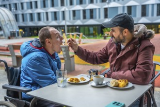 Man in wheelchair being fed by caregiver at an outdoor cafe table, showing compassion, inclusion