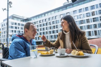 Woman feeding a smiling man with a disability, enjoying a meal and coffee at an outdoor cafe,
