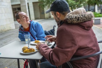 Two friends sharing an outdoor meal on a city terrace, both men are using wheelchairs and laughing,