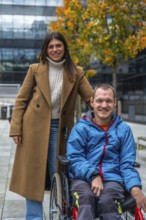 Young woman pushes her smiling partner in a wheelchair along an accessible urban pathway amid