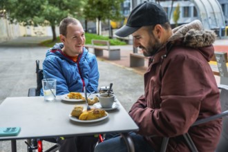 Young man with a disability smiling and relaxing in a wheelchair at an outdoor cafe table, enjoying