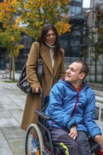 Cheerful caregiver standing behind a young man in a wheelchair, pushing him along an urban sidewalk