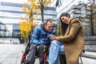 Man in a wheelchair and woman sitting on a bench, sharing a laugh and looking at a mobile phone