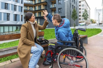 Female caregiver high fiving a young man in a wheelchair while sitting on a city park bench,