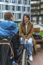 Young woman sitting on a park bench talking and smiling with a man in a wheelchair, showing