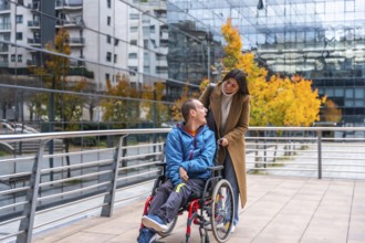 Caregiver pushing an adult man in a wheelchair on a modern city street, both individuals smiling