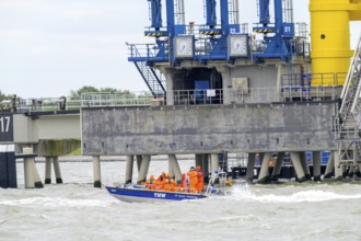 THW lifeboat with crew in orange clothing sails in front of port facilities on rough water,