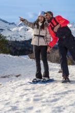 Smiling couple snowshoeing across snowy mountain slopes, pointing toward a breathtaking winter