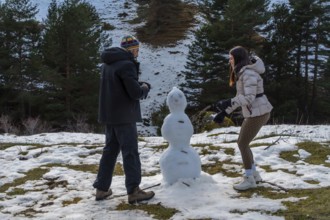 Couple building a snowman outdoors on a snowy hillside, wearing warm winter clothing and enjoying a