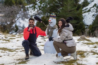 Smiling couple crouches beside a snowman in a snowy mountain forest, enjoying playful winter