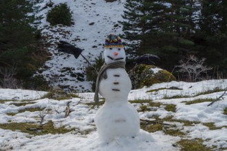 Snowman standing on a partially snow covered hillside, wearing a colorful knitted hat, grey scarf,