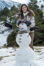 Young woman smiling while creating a snowman with a hat, scarf, carrot nose, and tree branch arms,