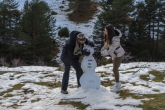 Young couple building a snowman in a snowy mountain pine forest, laughing and bonding outdoors on a