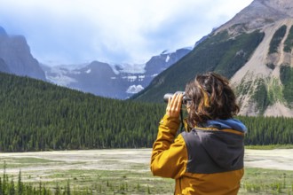 Female hiker wearing a yellow jacket using binoculars while observing a glacier in the canadian