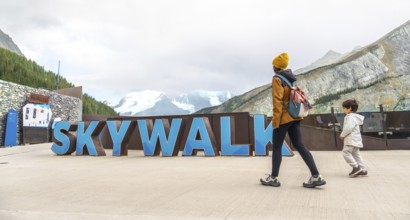 Tourists walking by large skywalk sign with athabasca glacier and mountains in background at