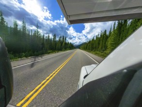 White vehicle driving along icefields parkway, surrounded by lush forests, under a bright blue sky