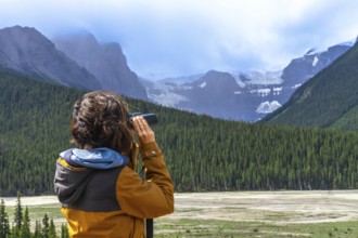 Tourist wearing an orange jacket uses binoculars to observe a glacier in the canadian rockies,