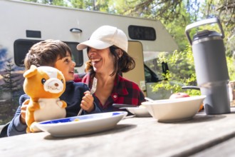 Mother and son laughing together while enjoying breakfast at a picnic table outside their camper
