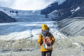 Female tourist in a yellow beanie and jacket, carrying a backpack, admiring the melting athabasca