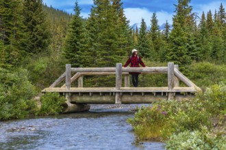 Female hiker wearing red plaid shirt and white hat is leaning on a wooden bridge railing over a