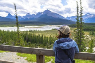 Tourist wearing a baseball cap and light blue jacket leaning on a wooden railing, taking in the