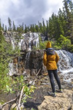 Female hiker wearing yellow beanie and jacket standing on rocks near waterfall and admiring