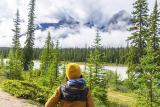 Hiker in a yellow beanie and jacket standing in a lush pine forest, admiring majestic mountain