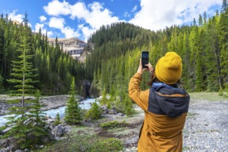 Hiker capturing smartphone images of a turquoise waterfall flowing through a lush pine forest along
