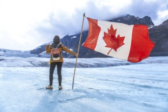 Female tourist is holding large canadian flag while standing on surface of athabasca glacier in