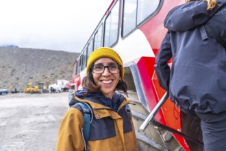 Tourist wearing a yellow beanie and brown jacket smiles near a large red bus at the athabasca