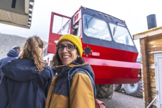 Tourist wearing a yellow beanie and glasses smiles near a large red bus, used to transport visitors