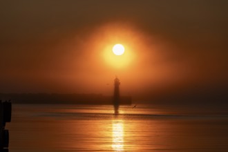 A lighthouse at the Vardö pier stands silhouetted against an orange sunset over the sea reflecting